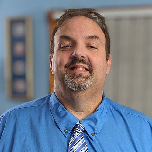 Man in a sky blue shirt and tie smiles for the camera.