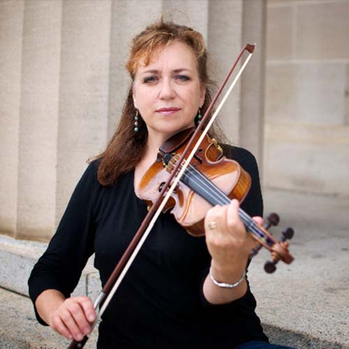 Woman standing outside in front of a marble pillar, holding a violin as if getting ready to play it.