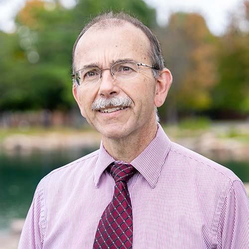 Man standing outside in the sunshine, wearing a pink dress shirt and maroon tie smiles at the camera