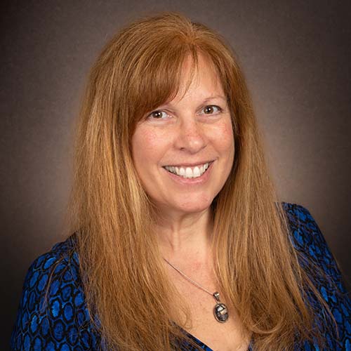 Woman with long strawberry blond hair and a blue v-neck dress shirt sits for a professional headshot