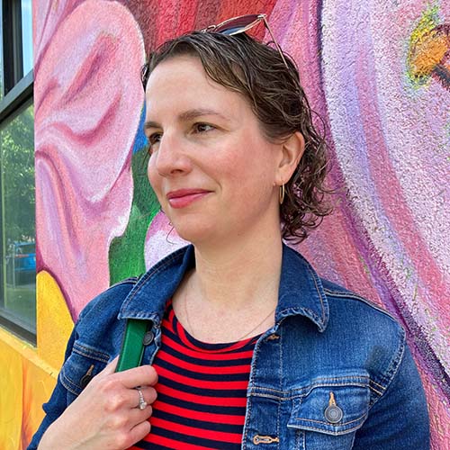 Woman with a striped shirt and jean jacket stands outside in front of a mural