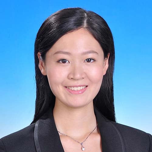 Woman with a dark suit jacket, long dark hair sits against a sky blue backdrop for a professional headshot
