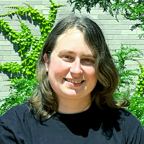 Woman with a black t-shirt stands outside on a sunny day in front of a brick building with ivy growing on it upwards