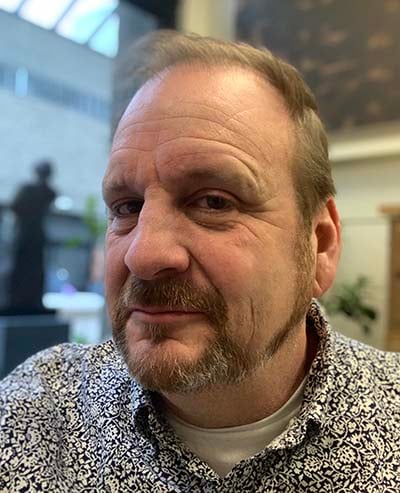 man with a coy smile, head tilted slightly with a wild pattern button-up shirt