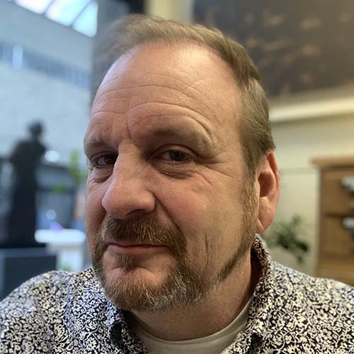 Man with a well-trimmed beard smirking, looking at camera with a slight side profile view. He's wearing a black and white flowery collared dress shirt.