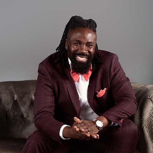 Black man sitting on a couch, wearing a dark plum suit and peach bow tie.