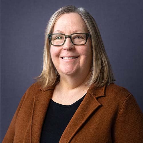 Woman with shoulder length dark blond hair wearing a brown suit jacket sits for a professional photo, smiling brightly.