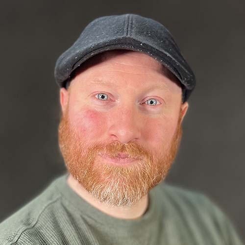 Man with a red beard and paperboy cap sits for a professional headshot