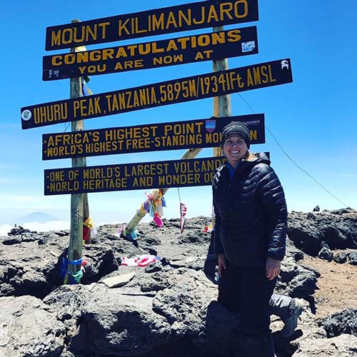 Woman in winter gear standing next to Mount Kilimanjara sign, atop Africa's highest point.