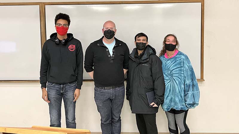 Three students and professor stand in front of a white board