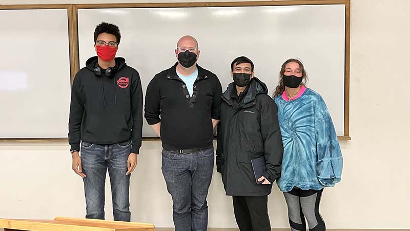 Three students and professor stand in front of a white board