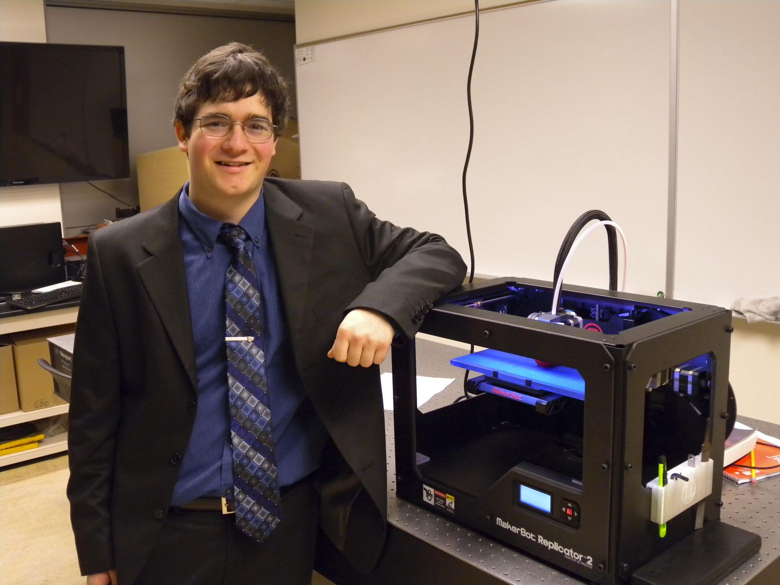 Justin is wearing a suit and tie, and standing next to physics equipment in a lab classroom