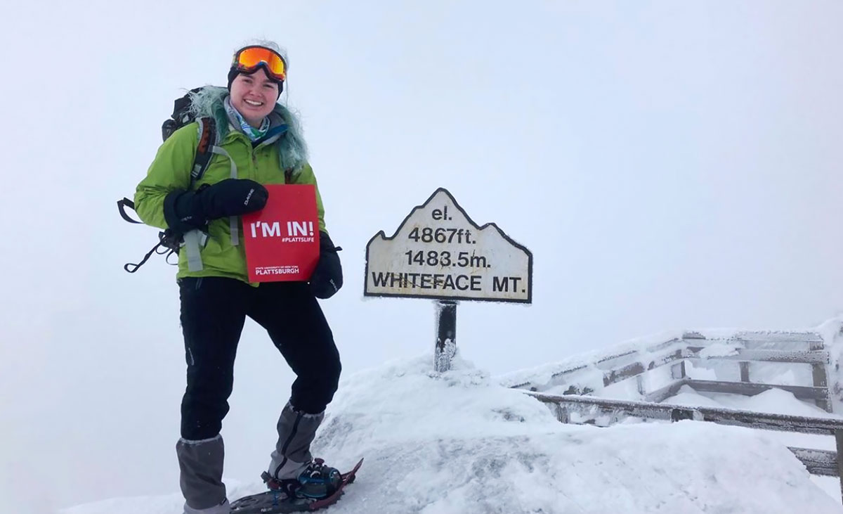 Katie Hale, dressed in snow gear and snowshoes, stands atop a snowdrift next to a Whiteface summit altitude marker holding up her acceptance packet Photo by @katie._hale