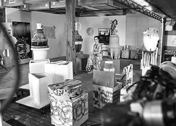Black and white image of the artist sitting in th emiddle of a rustic space, surrounded by large ceramic vessels