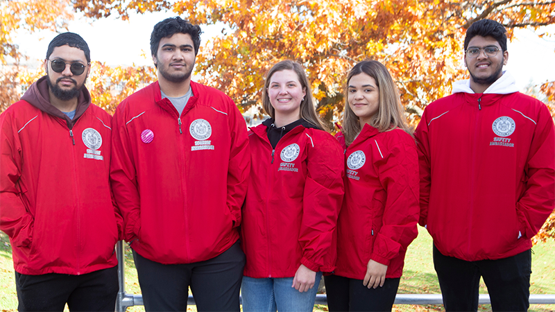 Five students with Safety Ambassador jackets on stand shoulder to shoulder in front of an orange-colored fall tree