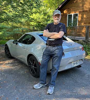 A man stands against a Mazda sports car with his arms crossed, smiling.