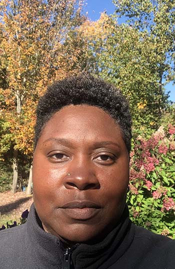 Selfie of artist, outside on a sunny fall day, surrounded by colored leaves on the trees.