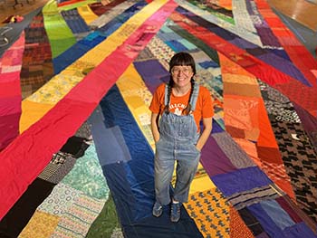 Portrait of Amanda Browder. She's standing on a giant quilted flag, she's wearing overalls and braided low pigtails.