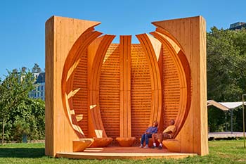 A large wooden structure almost resembling a pumpkin. Inside are benches on which a couple sit relaxing.